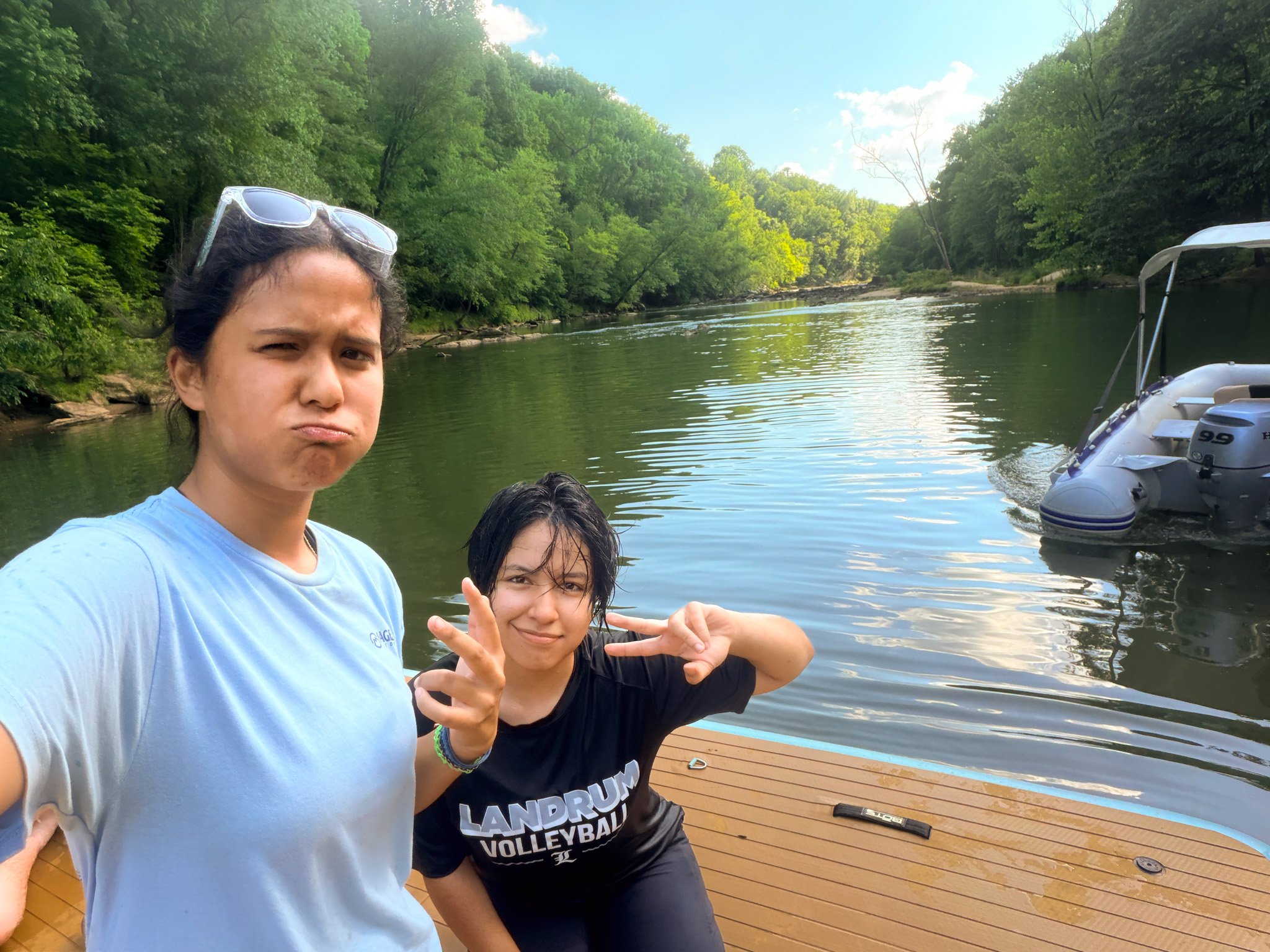 Two young girls posing by a river with boats and green trees, with one girl wearing goggles on her head and the other making a peace sign.