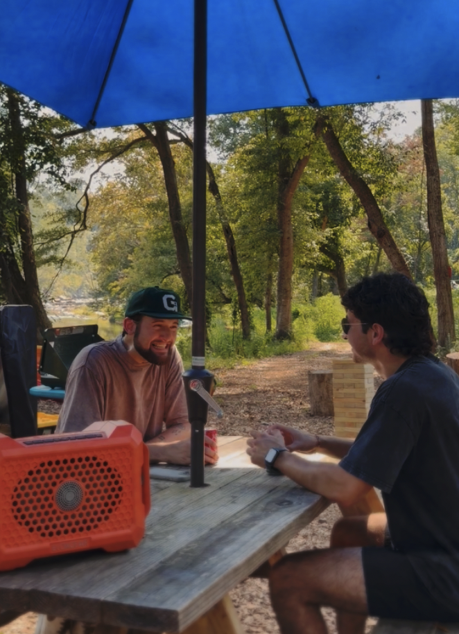 Two men sitting at a wooden picnic table outdoors, engaged in conversation under a large blue umbrella, with trees and a dirt path in the background.