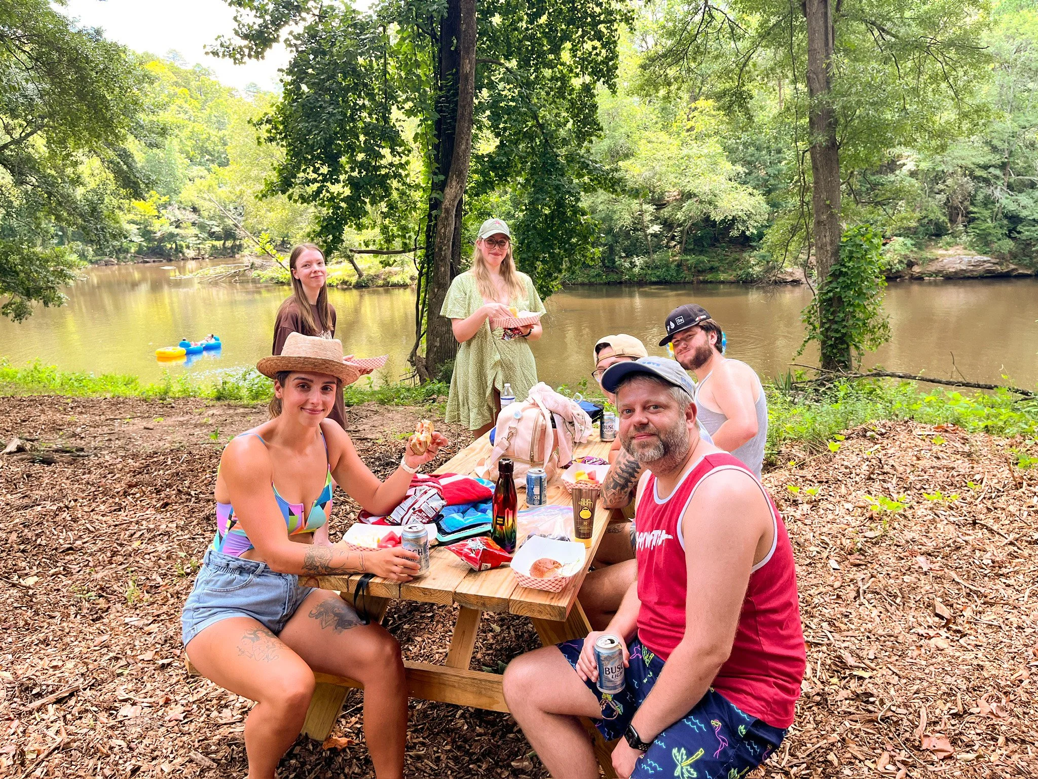 A group of seven friends having a picnic by a river in a wooded park, enjoying food and drinks on a wooden table.