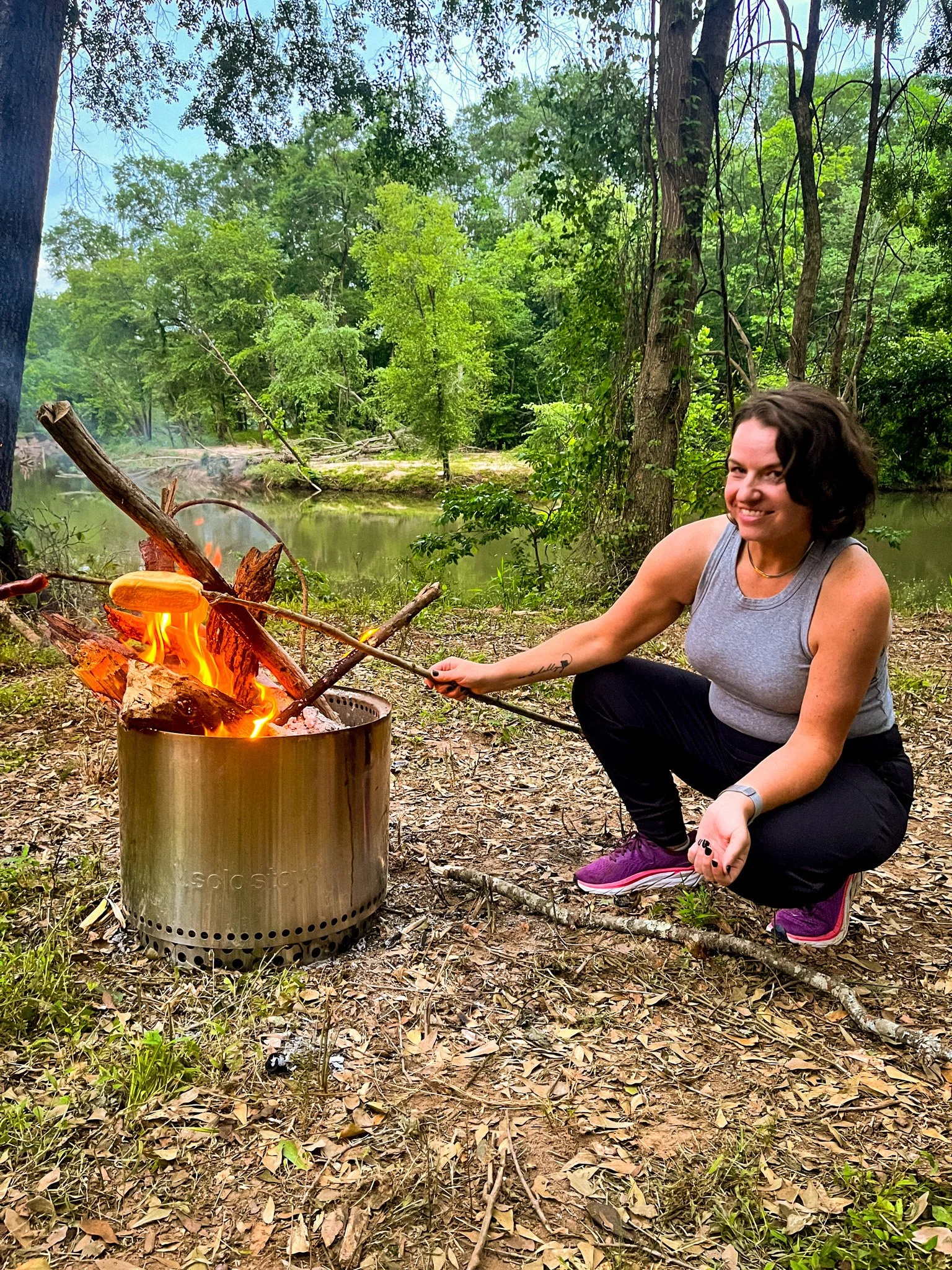 A woman with short dark hair smiling and squatting next to a campfire with flames, in a wooded area near a river surrounded by green trees.