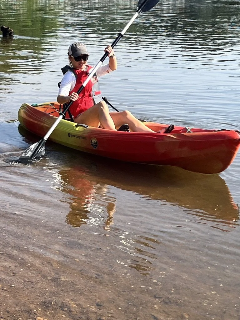 Person sitting in a red and yellow kayak on a body of water, holding a paddle with both hands, wearing sunglasses, a baseball cap, a white shirt, a red life jacket, and shorts.