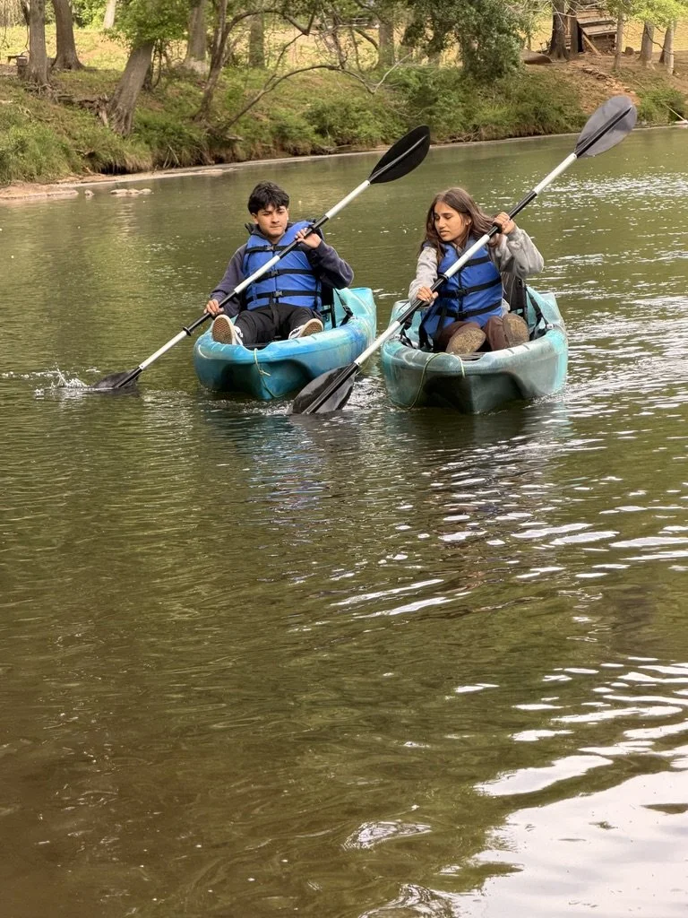 Two kids kayaking on a calm river surrounded by trees and a wooden staircase in the background, both wearing life jackets.