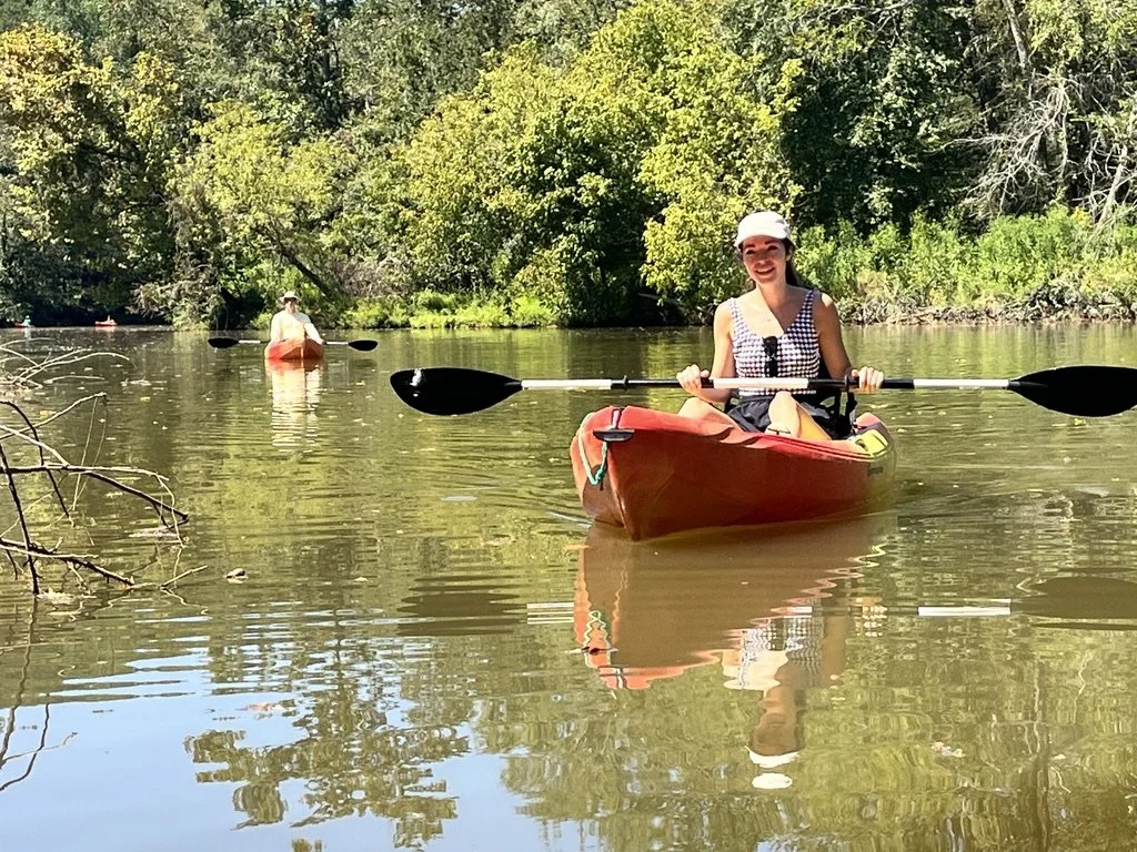 Two women kayaking on a calm river surrounded by green trees, with one woman in the foreground smiling and wearing a hat and sunglasses, and another in the background.