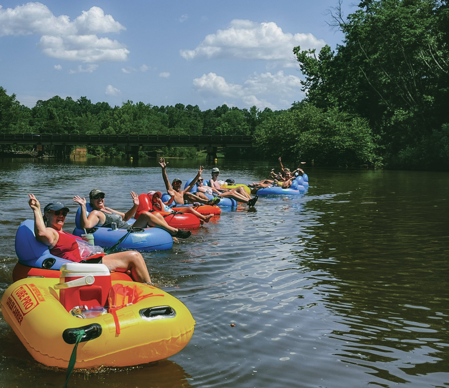 A group of people in colorful inner tubes floating on a river on a sunny day, with a bridge and green trees in the background.