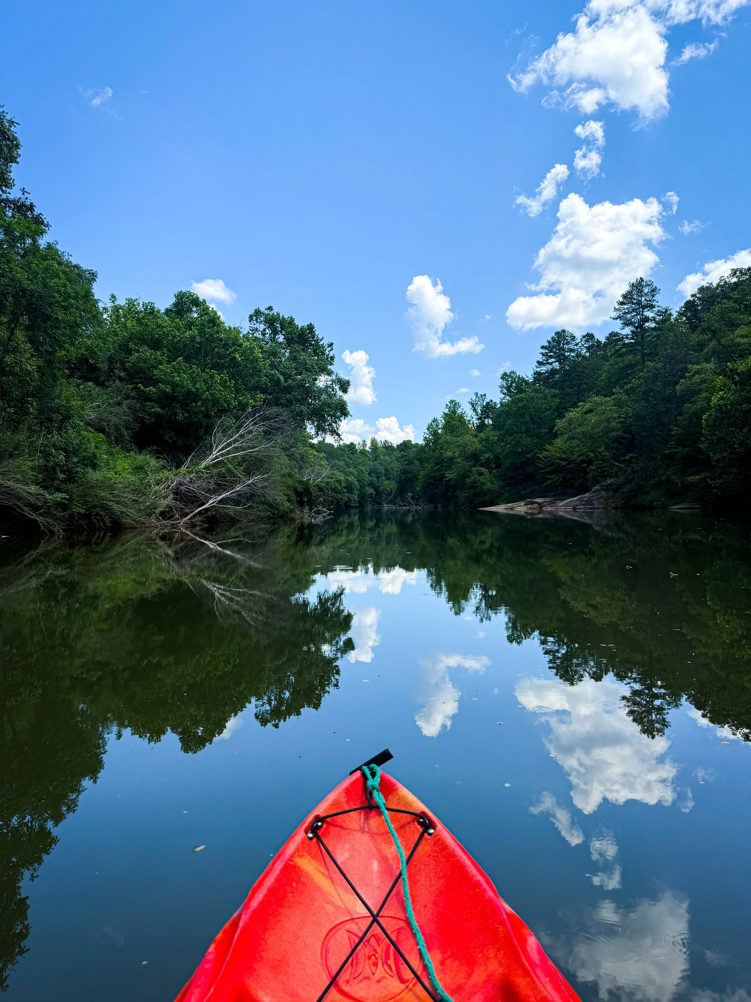 Red kayak on a calm river reflecting the blue sky with white clouds and green trees on either side.