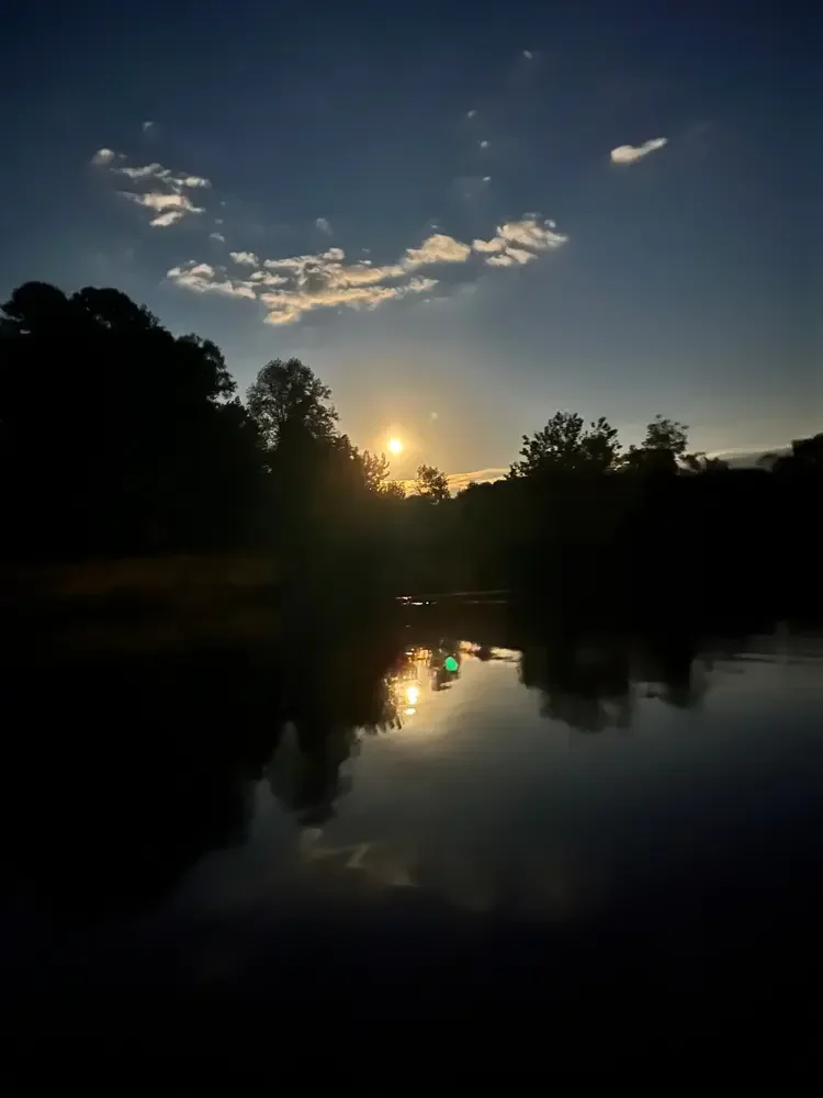 Sunset over a river with trees on both sides, partly cloudy sky, and the sun's reflection on the water.