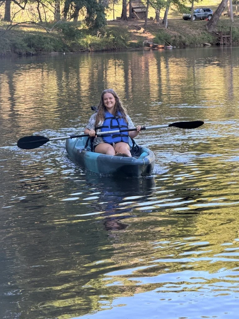 Young woman in a kayak on a calm river, smiling, holding a paddle, wearing a blue life jacket and casual clothing, with trees and cars in the background.