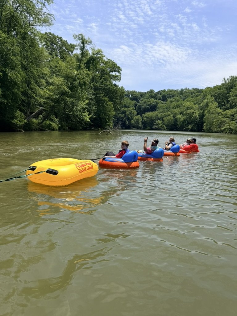 Four people riding in small boats on a river, surrounded by green trees under a partly cloudy sky.