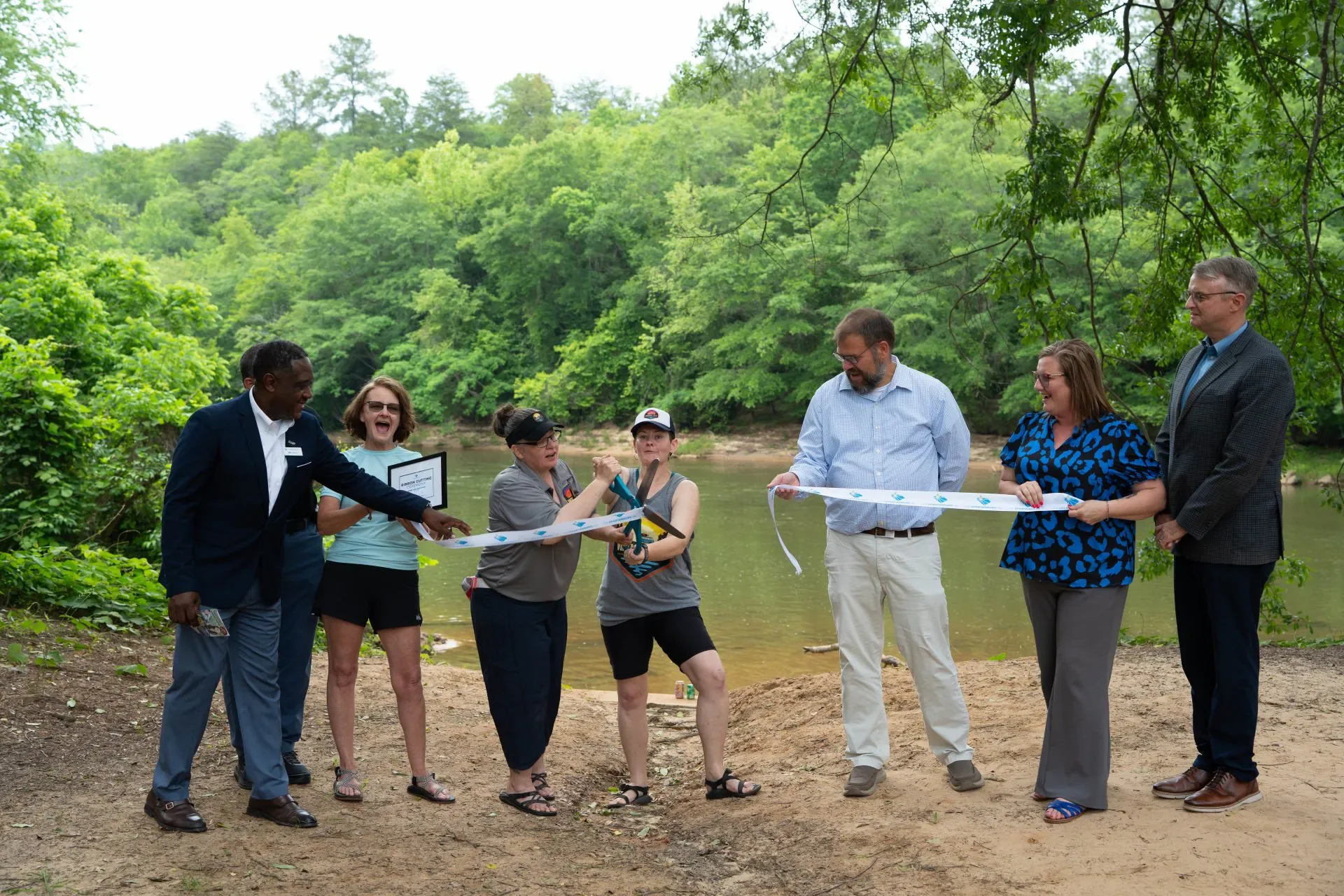 Group of people gathered for a ribbon-cutting ceremony by a river, with a green forest in the background.