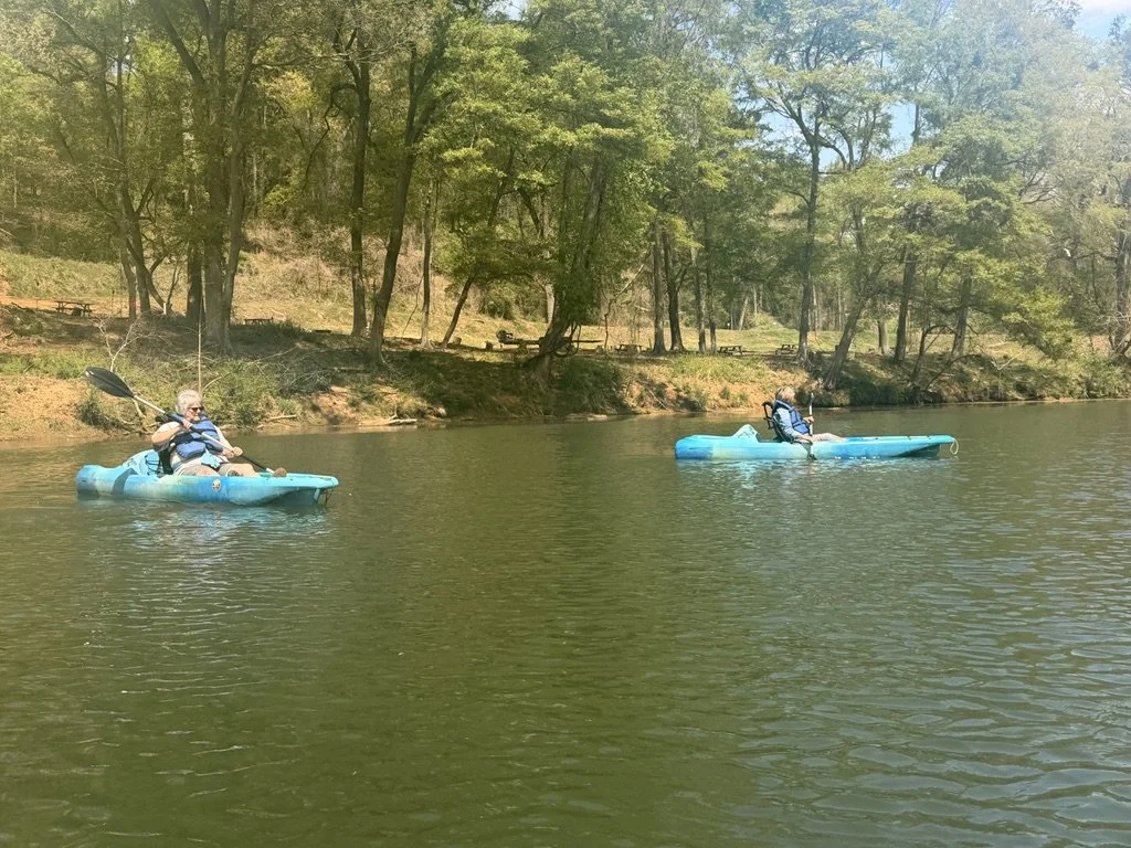 Two people kayaking on a calm lake surrounded by trees under a clear sky.