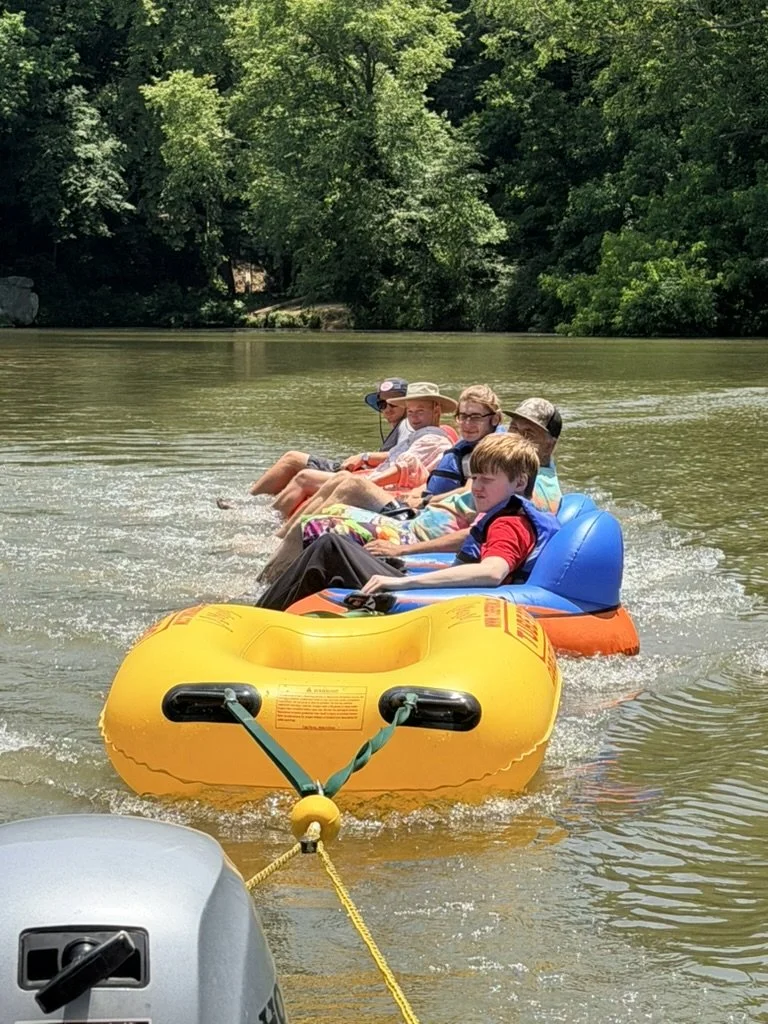 Six people sitting in a large inflatable boat being pulled by a motorboat on a river, surrounded by lush green trees.