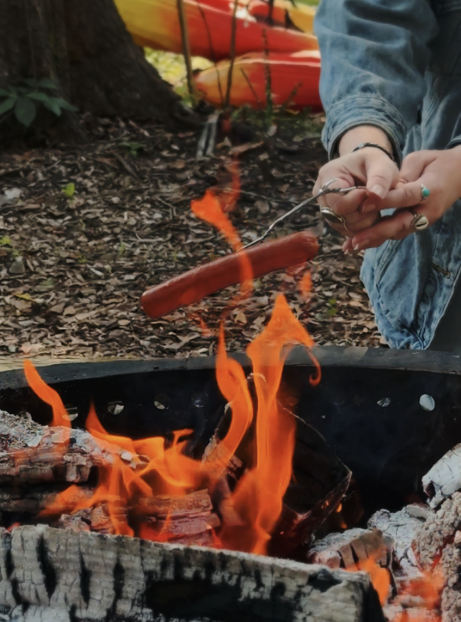 Person roasting a hot dog over an outdoor campfire in a forest setting.