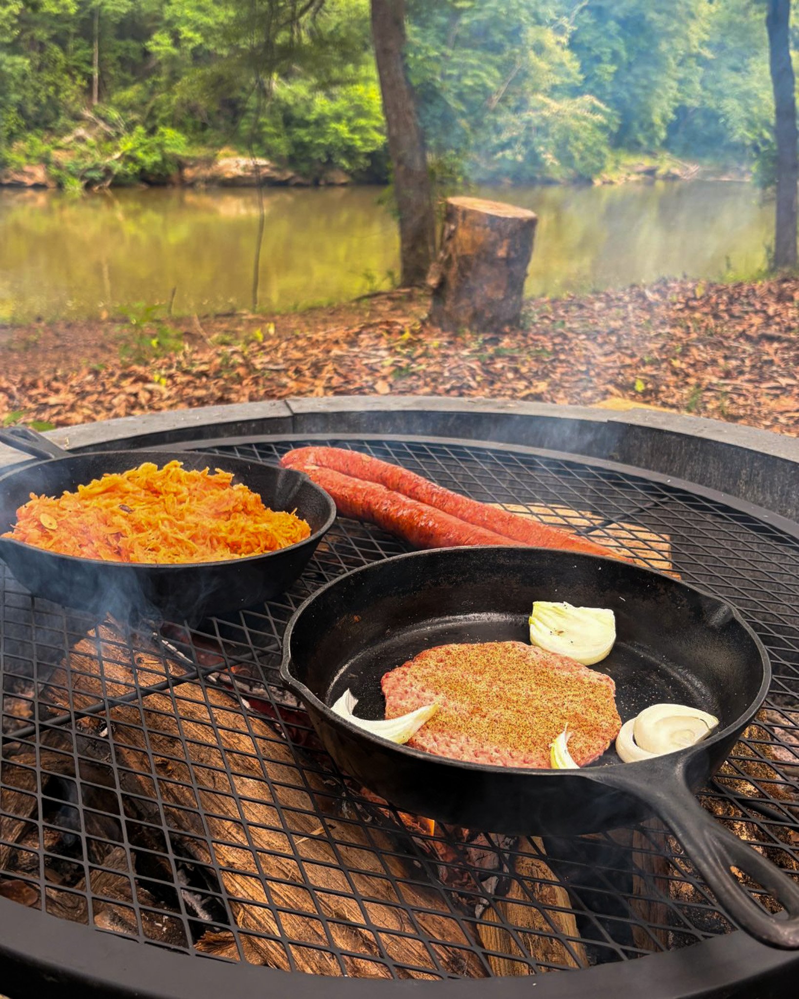 Cooking food on a grill outdoors near a river with trees and fallen leaves.