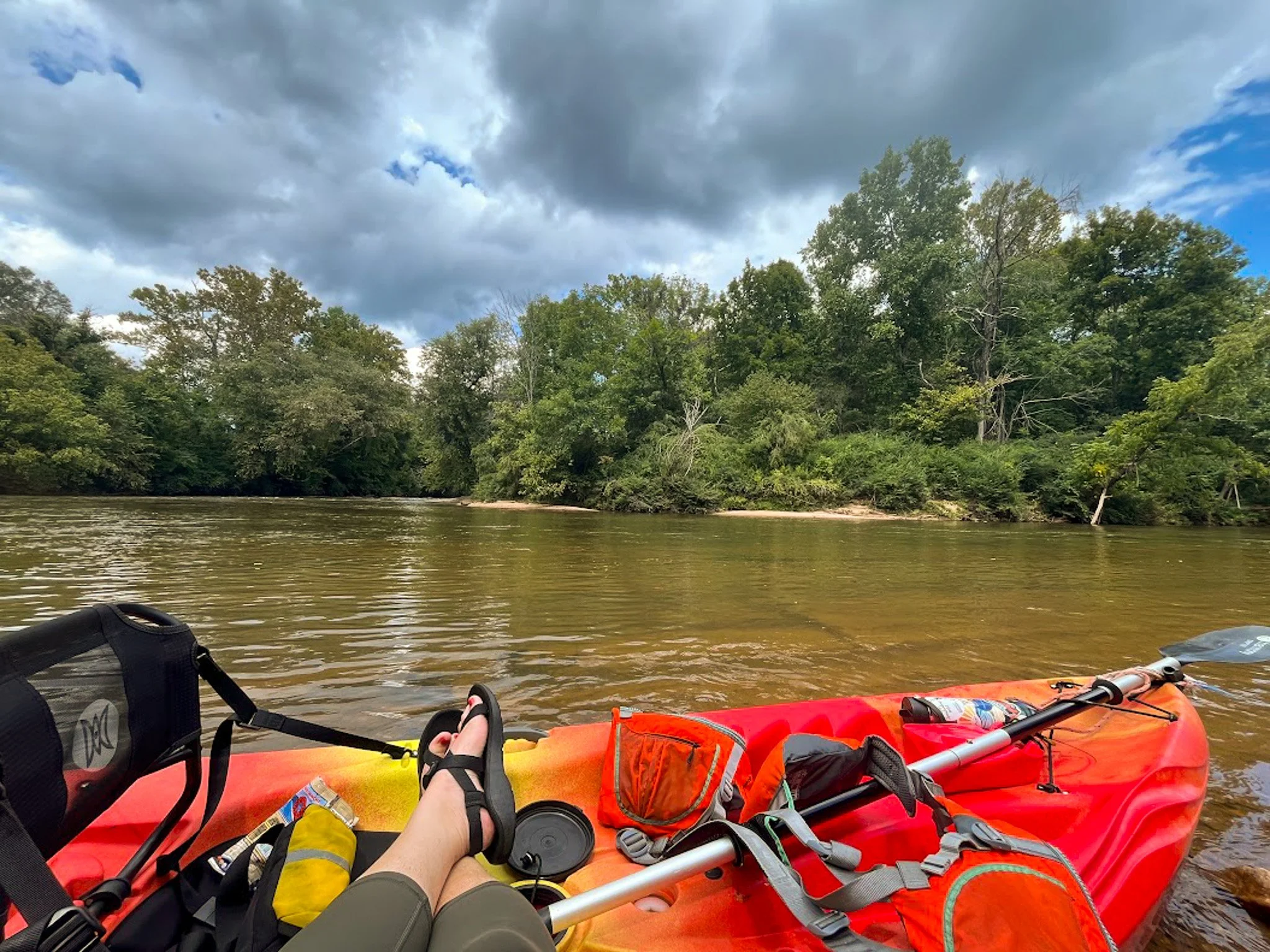View from a kayak on a river, showing the kayak's seating and gear, with lush green trees and a partly cloudy sky in the background.