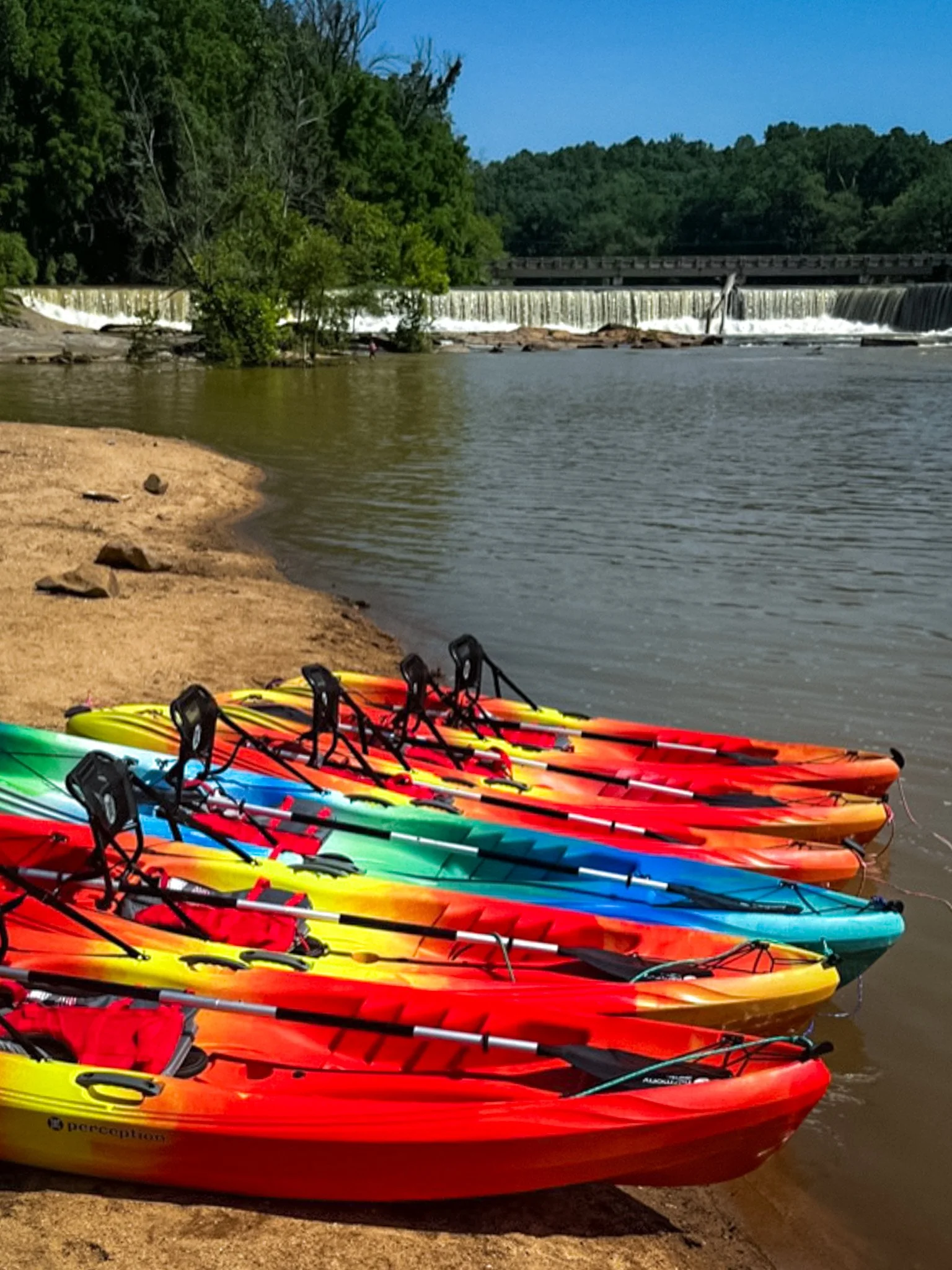 Multiple colorful kayaks lined up on the sandy shore of a river, with a waterfall and trees in the background under a clear blue sky.