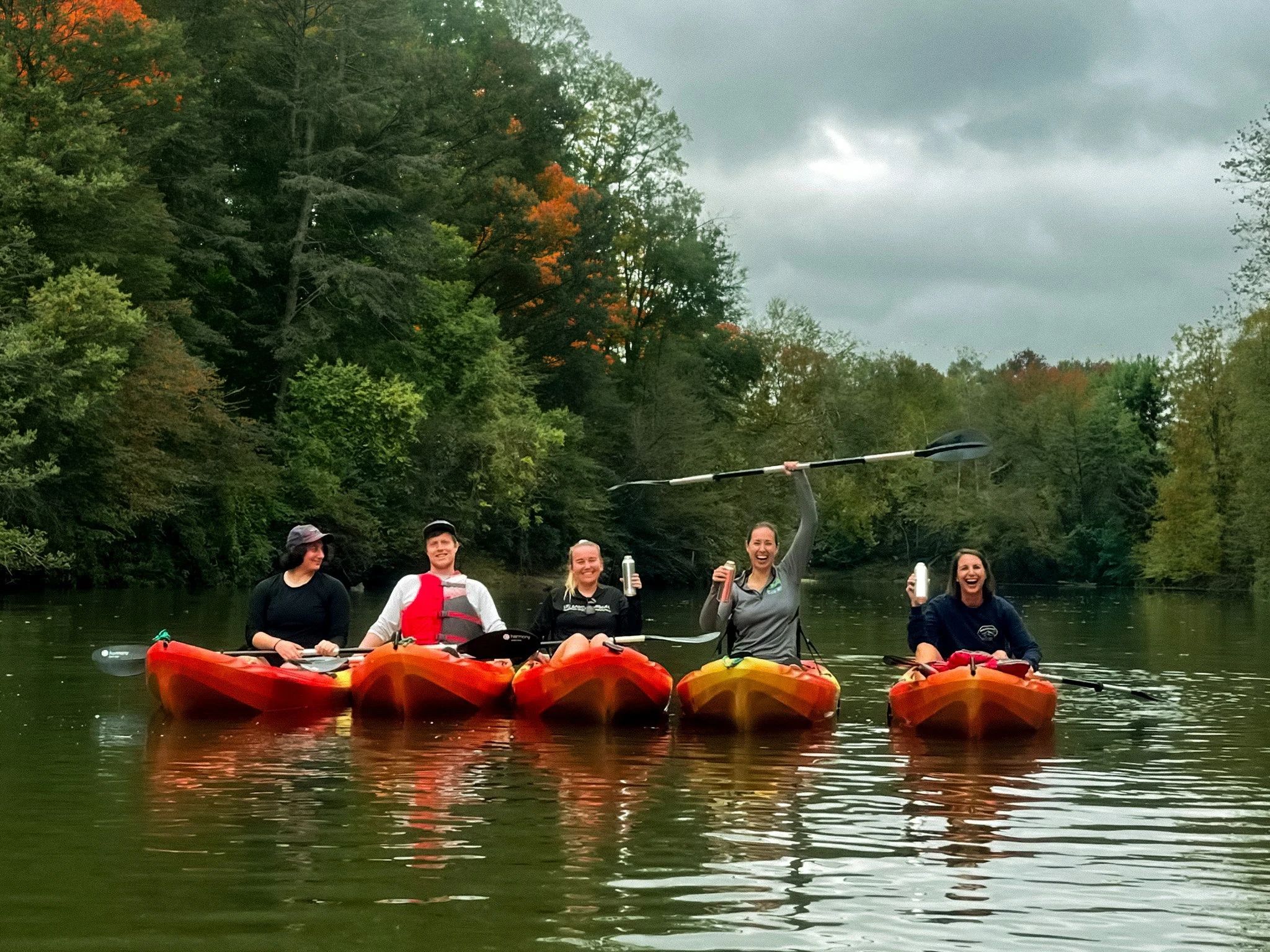 A group of five people in a kayak on a calm river, surrounded by lush green trees with some fall colors, under a cloudy sky.