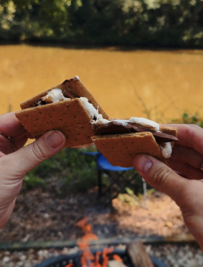 Close-up of a person holding a s'more in front of an outdoor campfire with a yellow river and green trees in the background.