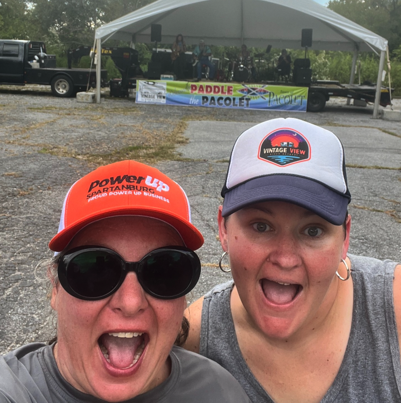 Two women taking a selfie outdoors in front of a stage with a band playing, under a white canopy. One woman wears sunglasses and a red cap with 'Power Up' logo, and the other a gray and black cap with 'Vintage View' logo. Behind them, a banner reads 'Paddle the PACELOT' and there are vehicles parked on a gravel lot.