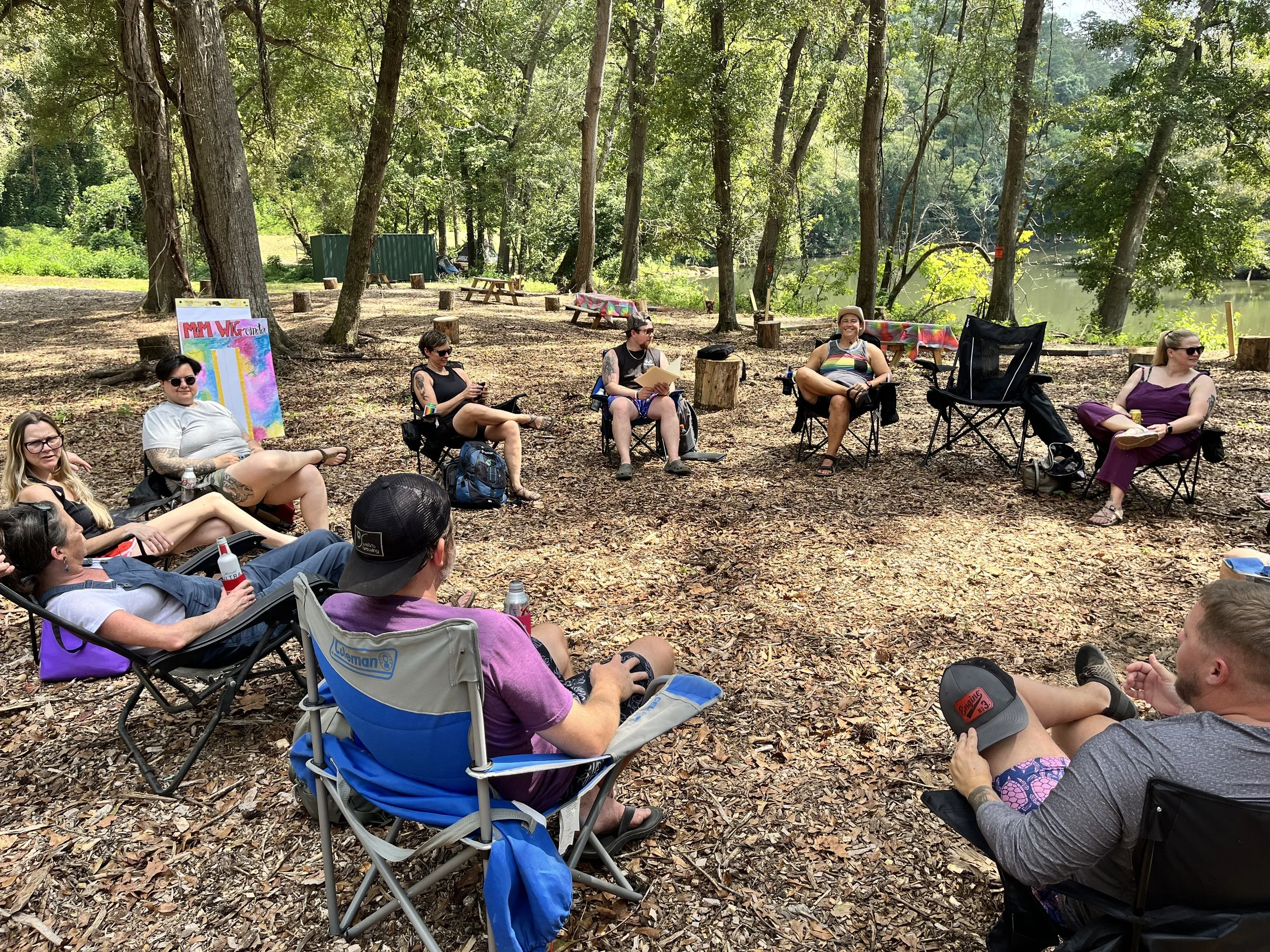 Group of people sitting in a circle on camping chairs in a wooded area near a river. Some are using their phones, and there are visible backpacks, a sign, and a table with a colorful cloth.