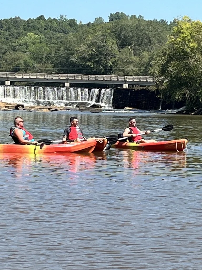 Three people kayaking on a river with a small waterfall in the background, surrounded by trees.