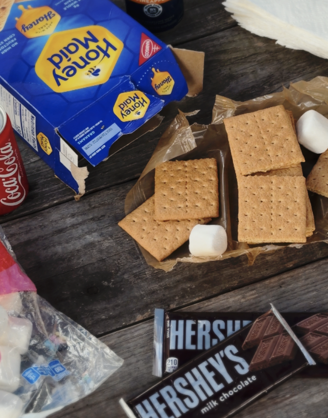 A wooden table with a partially opened box of Kraft Cheddar Macaroni & Cheese, a Hershey's milk chocolate bar, a Coca-Cola can, some marshmallows, graham crackers, and other snack items.