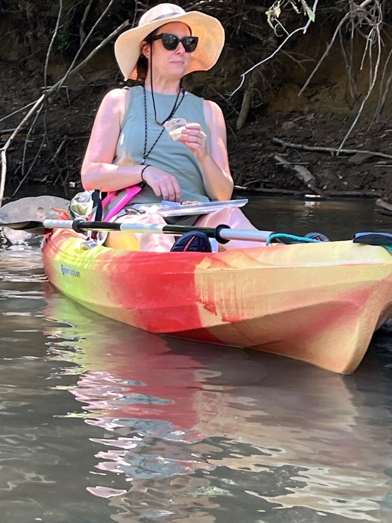 Woman wearing a sunhat, sunglasses, and a tank top sitting in a colorful kayak on a river, holding a snack and a clipboard.