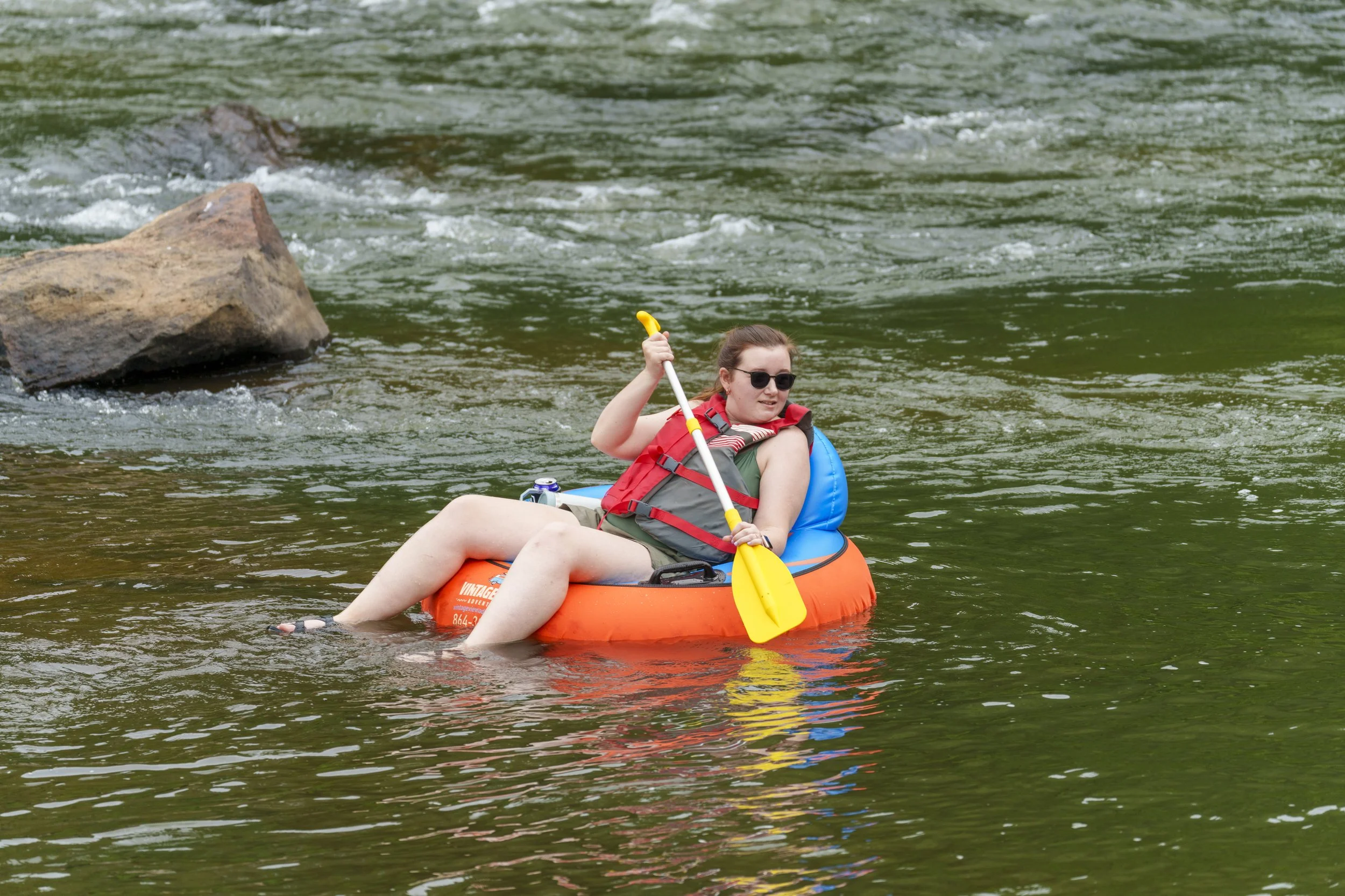 A person floating on a river in an orange kayak, wearing a red life jacket, sunglasses, and holding a yellow paddle.