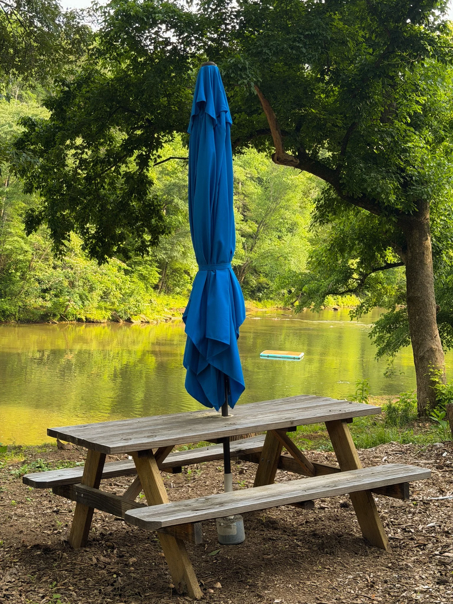 A closed blue patio umbrella on a wooden picnic table near a river, surrounded by lush green trees and foliage.
