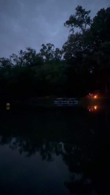 Nighttime scene of a dark lake with trees silhouetted against a cloudy sky, and a small campfire reflecting on the water.