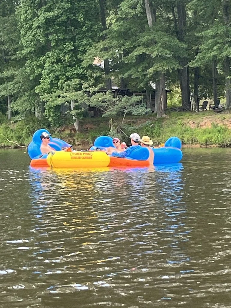 Group of people relaxing on an inflatable boat with blue cushions on a lake, surrounded by trees.