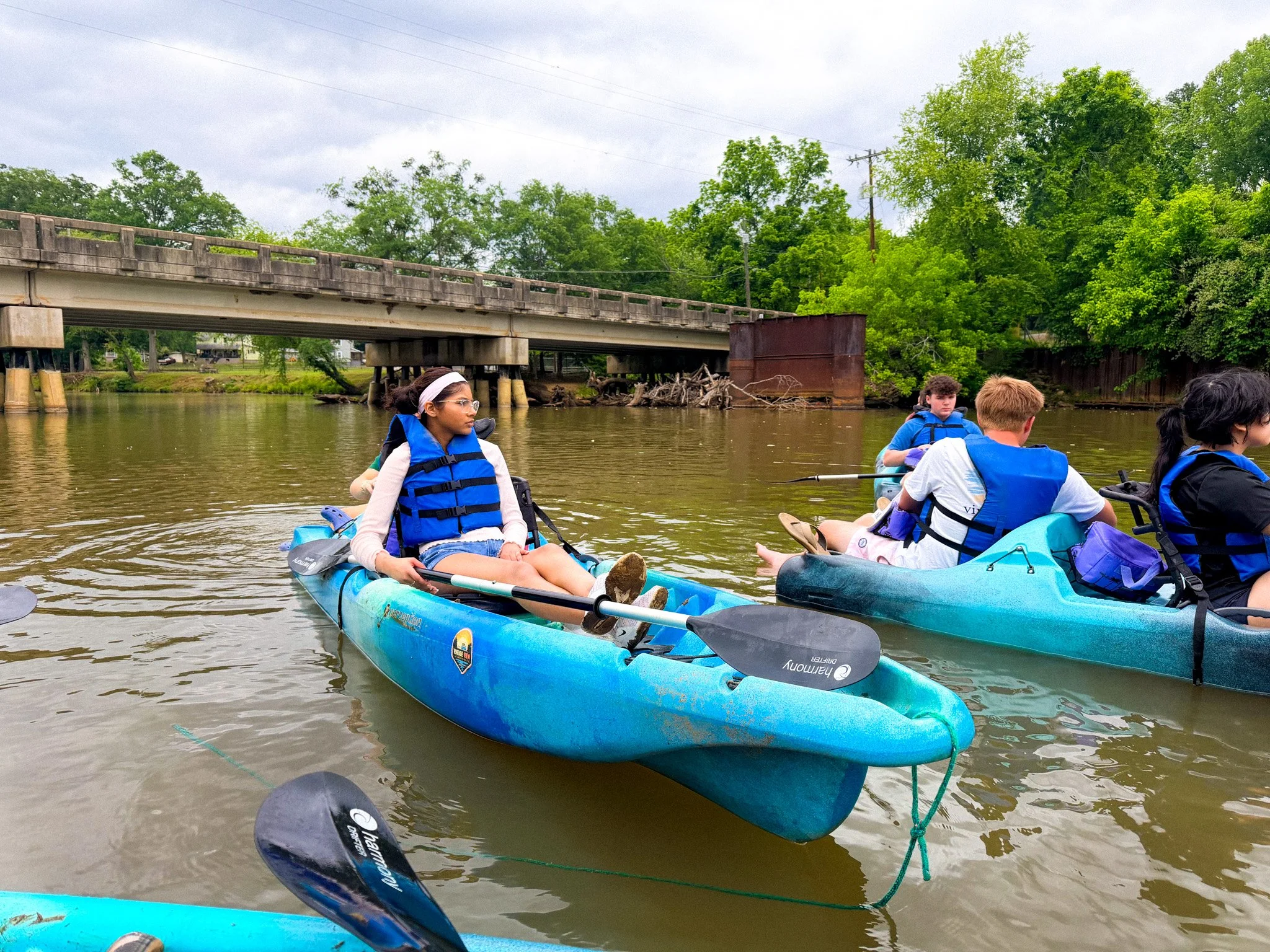 People kayak on a river near a bridge and green trees.