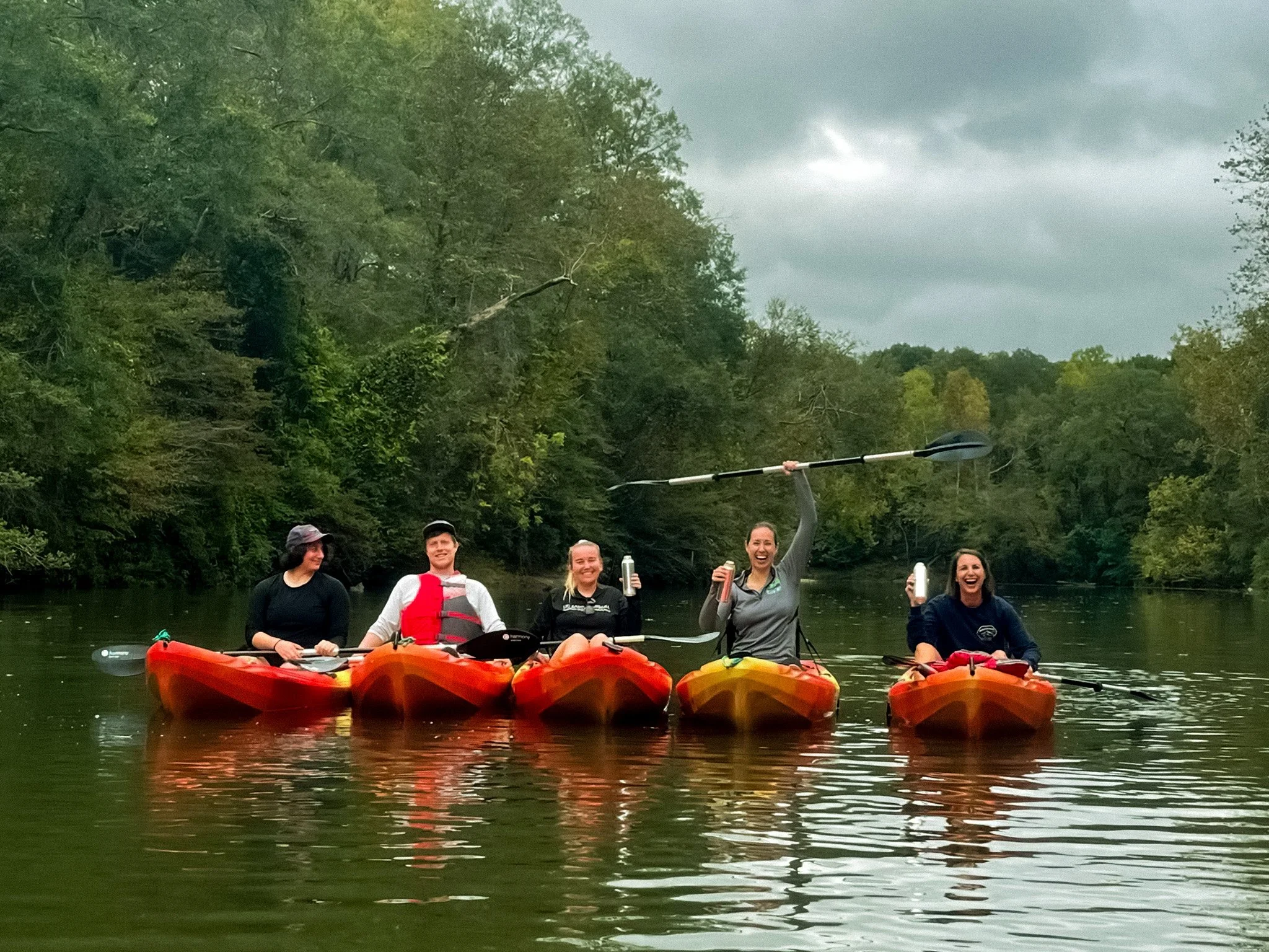 Group of five people kayaking on a river surrounded by trees, one person is holding a paddle aloft and smiling.