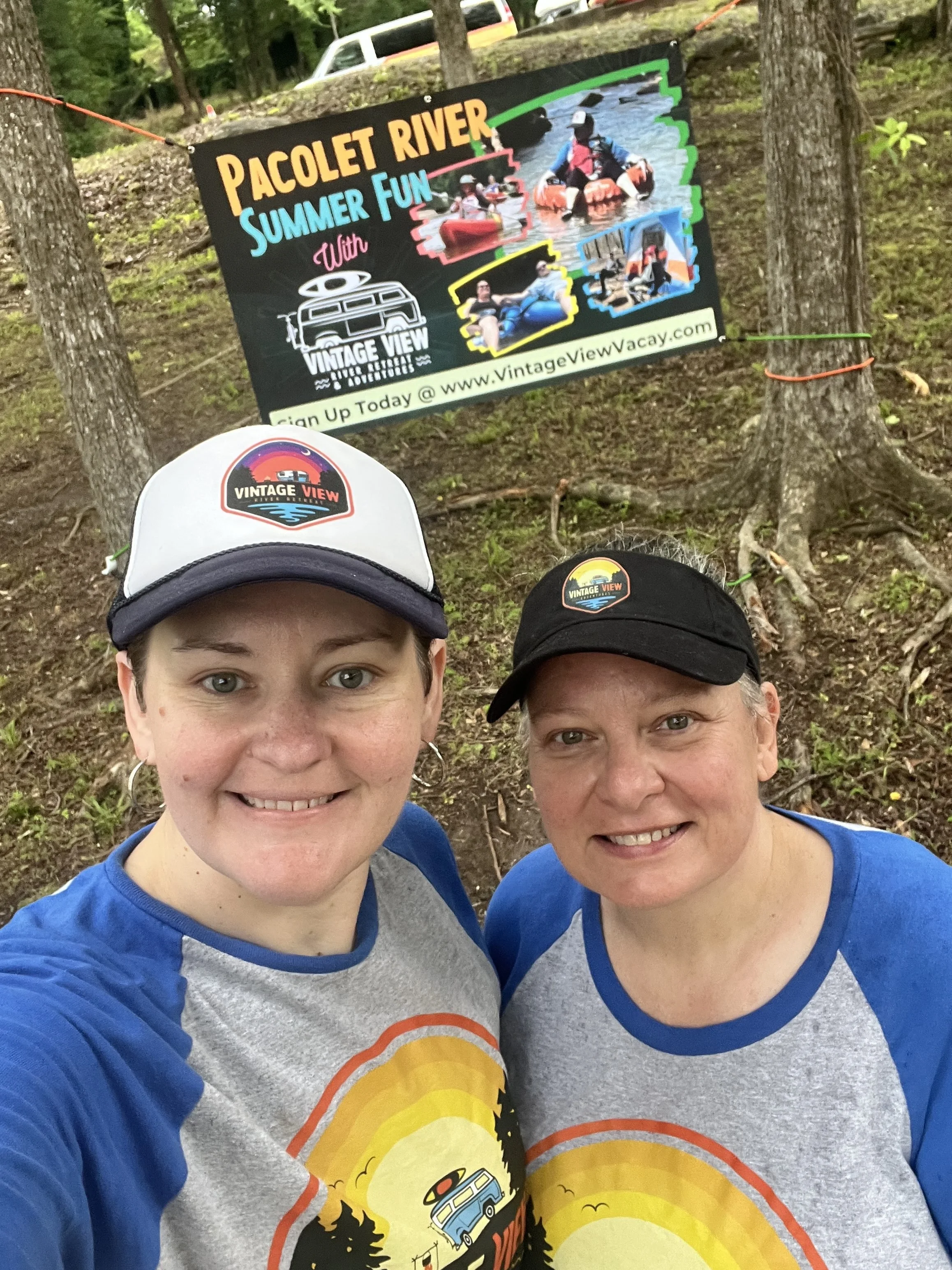 A selfie of two smiling women wearing gray and blue shirts with a vintage view logo on their hats. They are outdoors in a wooded area with a sign for Paolet River Summer Fun and a photo of people kayaking in the water in the background.
