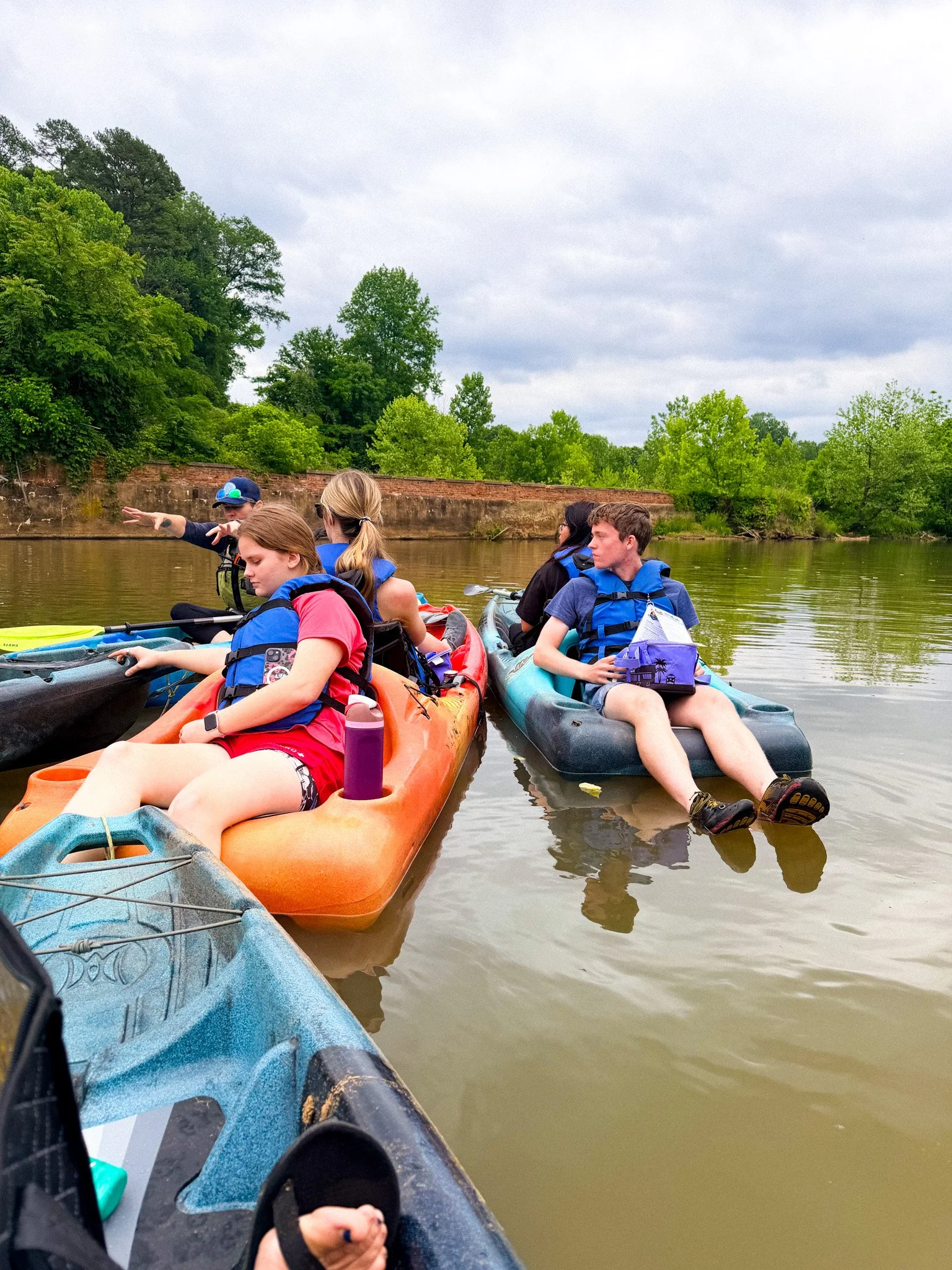 Group of people kayaking on a calm river with lush green trees and a brick wall in the background under a cloudy sky.