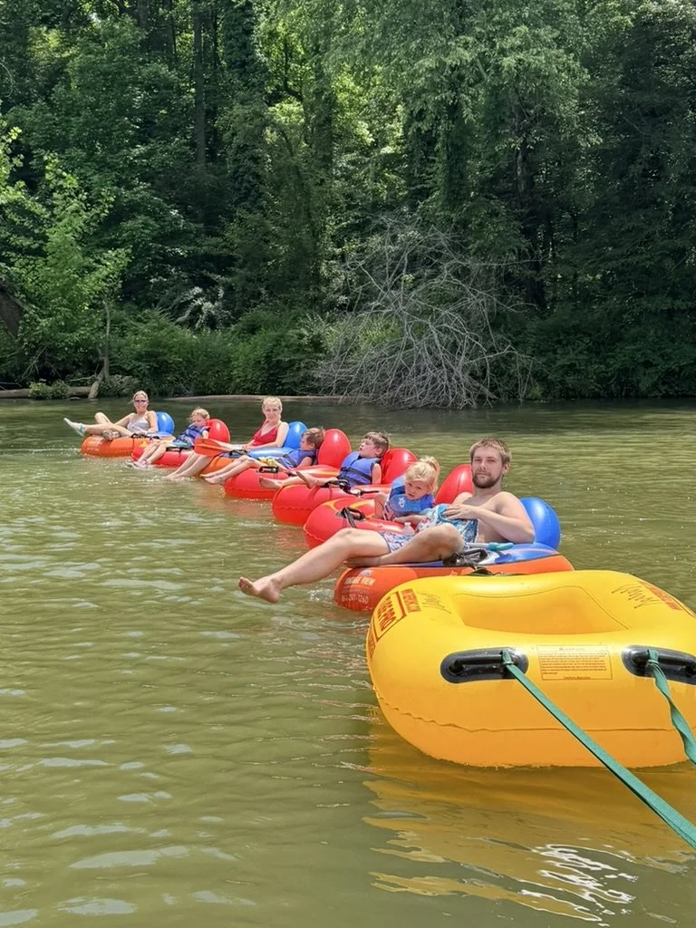 A group of people, including children and adults, relaxing on interconnected floating tubes in a river surrounded by lush green trees.
