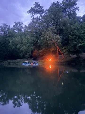 Calm river scene at dusk with a campfire near the shoreline surrounded by trees