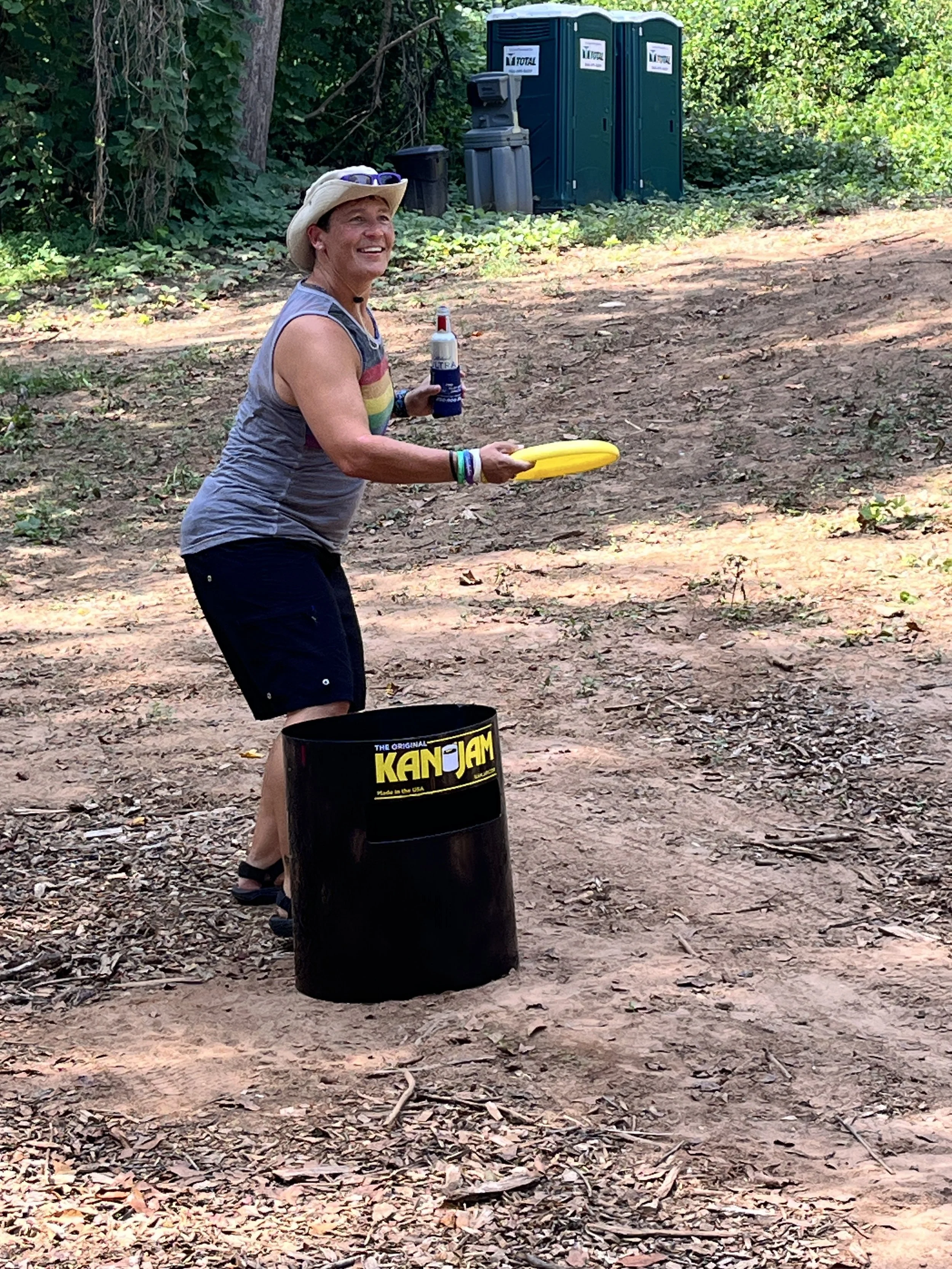 A woman in a sleeveless rainbow-colored top and shorts, wearing a wide brim hat and bracelets, smiling and holding a frisbee. She is standing outdoors in a wooded area with a black trash can in front of her and a barrel. In the background, there are 