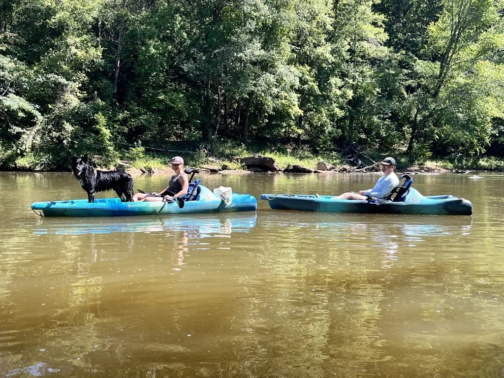 Two people in kayaks on a calm river with trees in the background. One person has a black dog in their kayak.