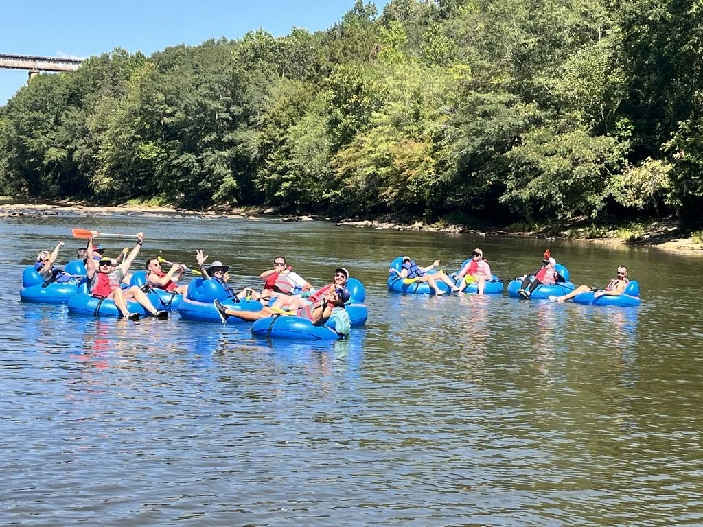 Group of people floating on the river in blue inflatable tubes, enjoying a sunny day with green trees and a bridge in the background.