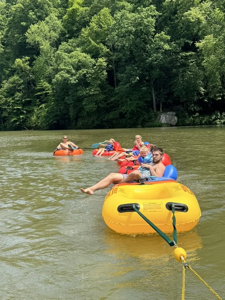 A group of people, including children and adults, floating on the water in inflatable tube rafts, with lush green trees in the background.