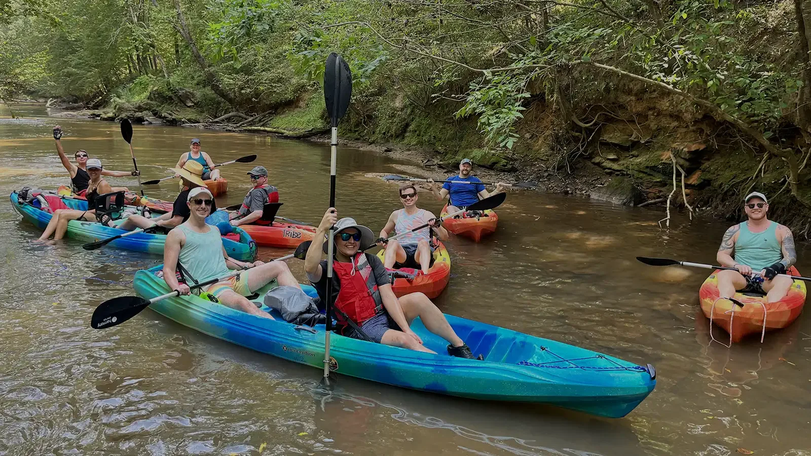 Group of people kayaking on a river surrounded by trees, some smiling and waving at the camera.