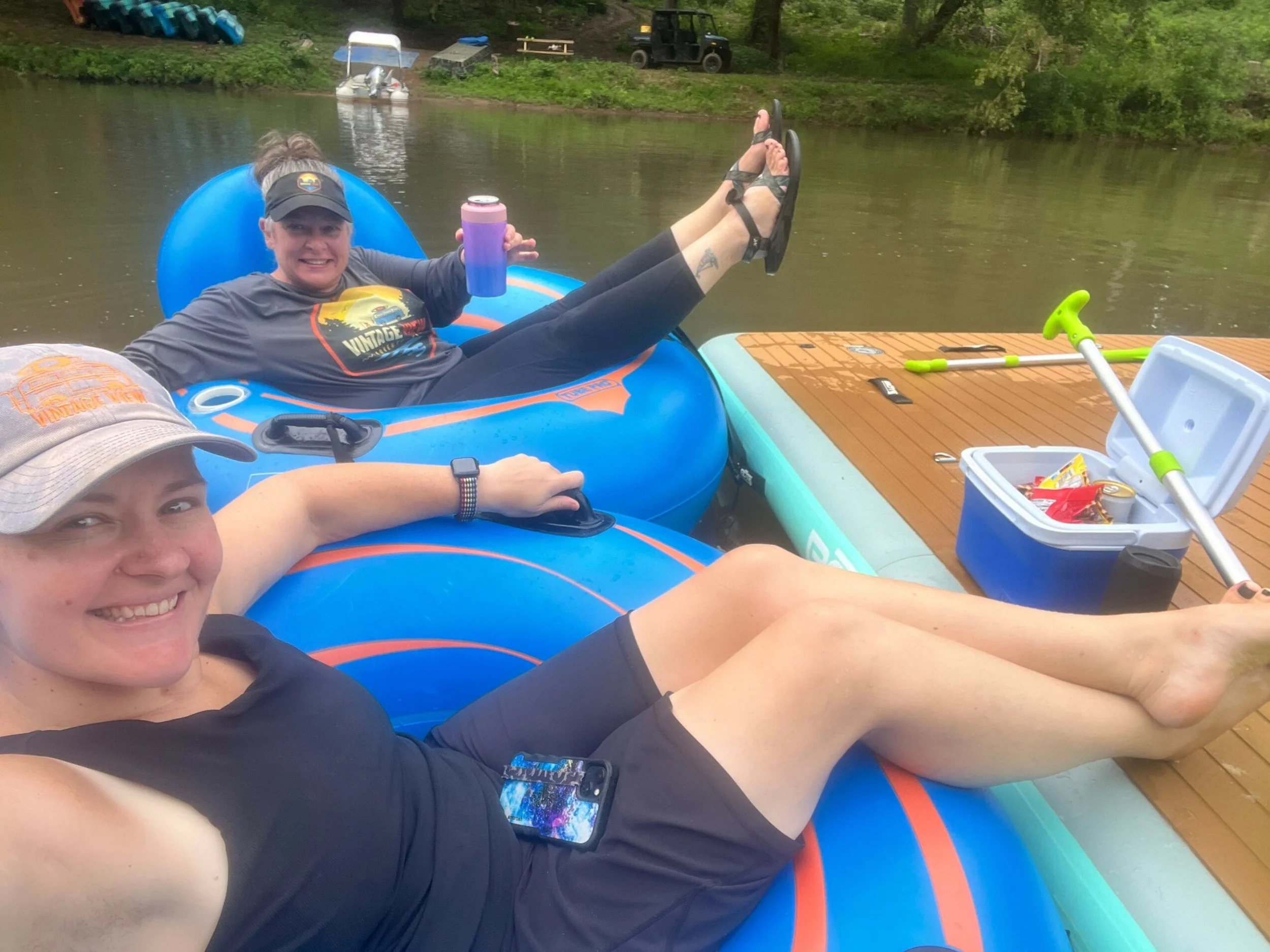 Two women and one man relaxing on an inflatable boat in a lake, smiling at the camera. One woman holds a drink, and the boat is next to a floating dock with various items and trees in the background.