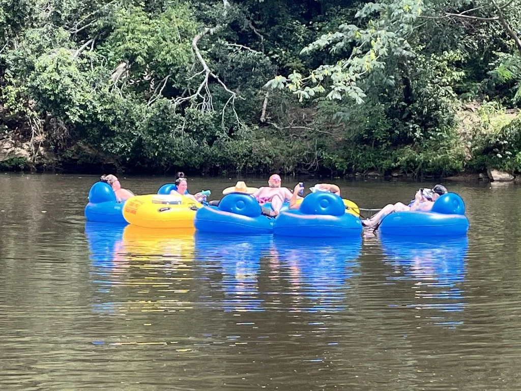 Five people relaxing on a large, inflatable water float on a river, with trees and greenery in the background.