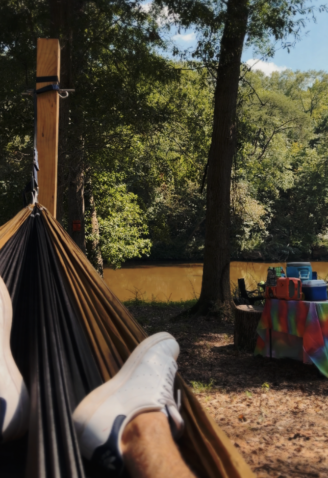 Person relaxing in a hammock near a river with trees and a table with supplies nearby.