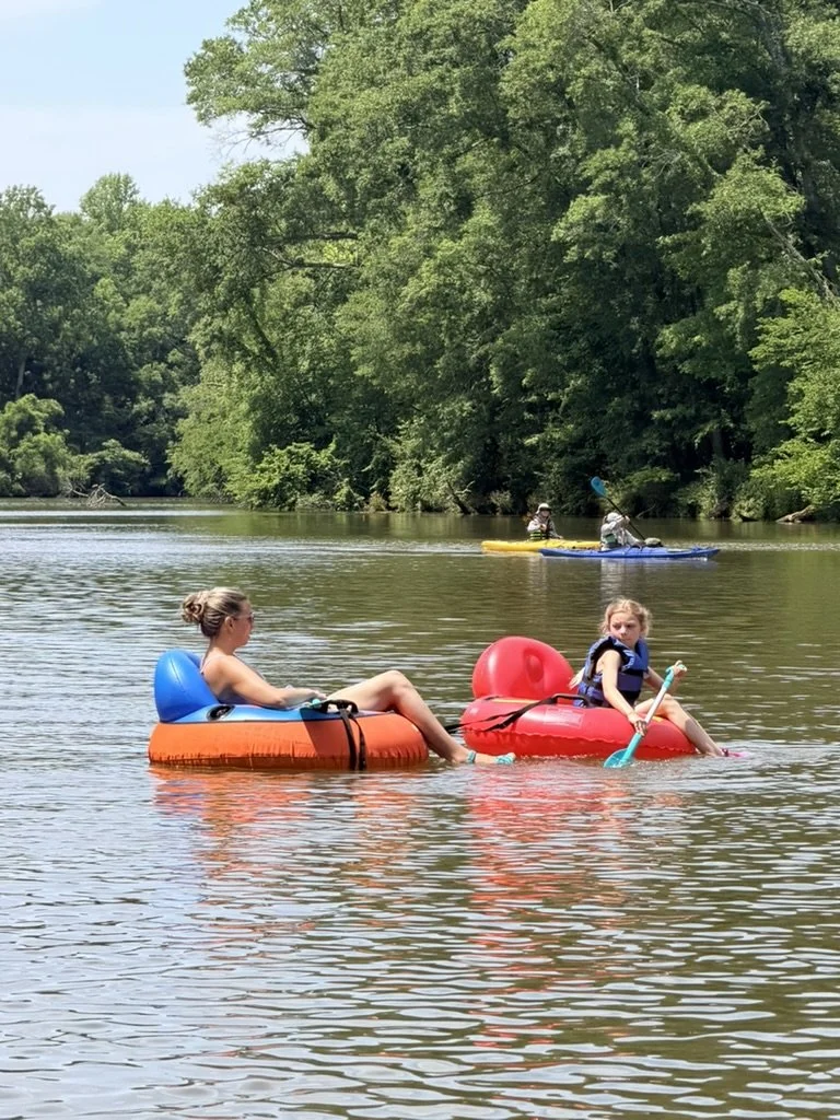 Two young girls relaxing on inflatable tubes in a calm river with lush green trees in the background, while two people kayak further upstream.