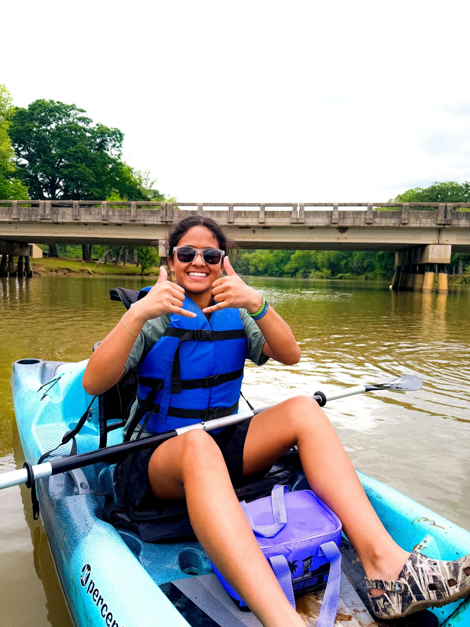 Woman smiling and giving thumbs up while sitting in a kayak on a river, with a bridge and green trees in the background.