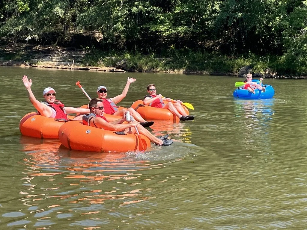 Four people floating on a river in orange inner tubes, with a fifth person floating on a blue inflatable ring in the background. The group appears to be enjoying a day of tubing, with trees lining the riverbank behind them.