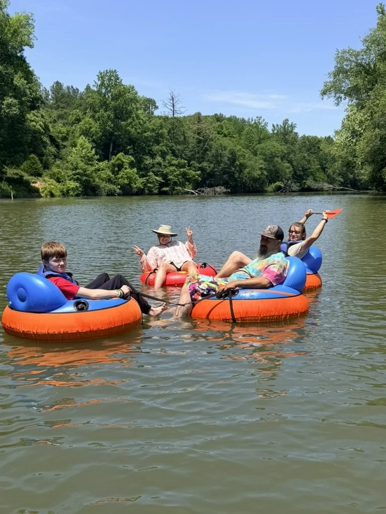 Four people on inflatable inner tubes floating on a calm river, surrounded by green trees under a blue sky.