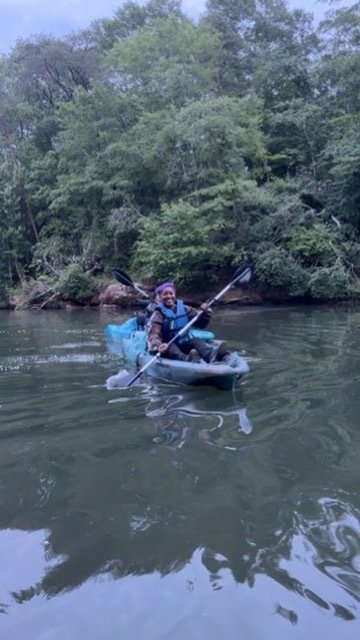 Person kayaking on a river surrounded by trees, wearing a life jacket and smiling.