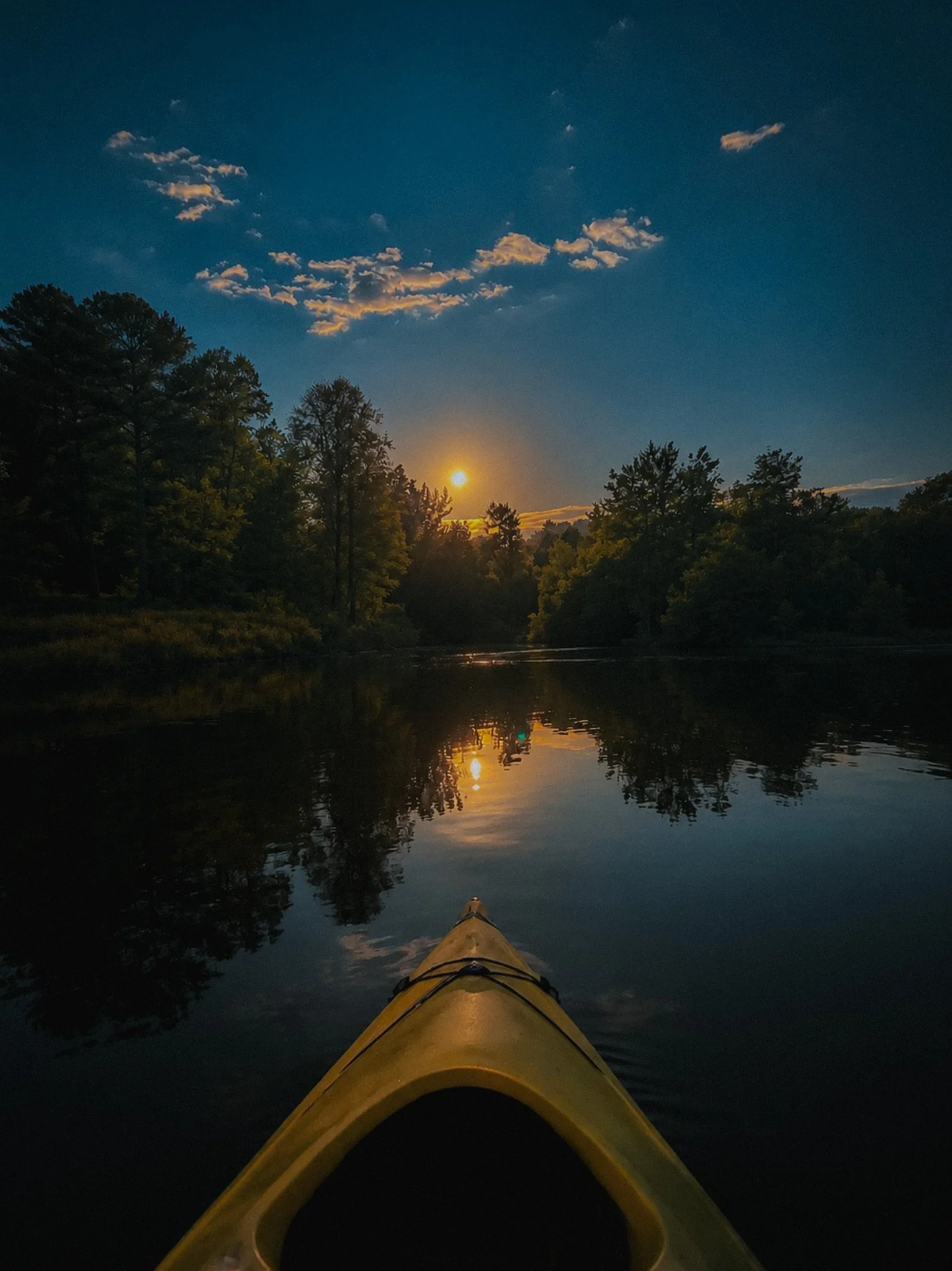 The photo shows the front view of a yellow kayak on a calm river or lake during sunset or sunrise. The sky is darkening with some clouds illuminated in the distance, and the silhouette of trees is visible along the shoreline.